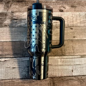 May include: A stainless steel Stanley tumbler with a black handle and lid, featuring a laser-etched design of a fishing rod, a large fish, and a vertical striped pattern with stars. The brand name Stanley is printed in black on the upper front.