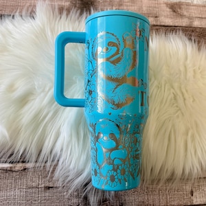 May include: A turquoise insulated tumbler with a handle, featuring a gold illustration of sloths and floral designs. The tumbler has a turquoise lid and handle. The design includes two sloths and the word "I".