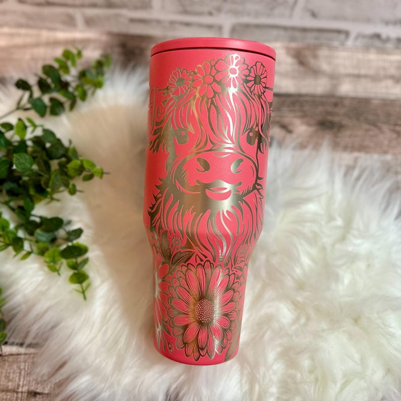 May include: A coral-colored tumbler with a floral and Highland cow design. The design is in a metallic gold color. The tumbler has a lid and is made of stainless steel. The tumbler is a great gift for anyone who loves cows.