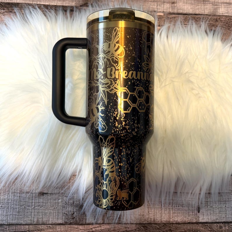 May include: A black and gold insulated tumbler with a handle. The tumbler features a bee and honeycomb design with the name "Ms. Breanna" in gold lettering. The lid is gold and the handle is black.