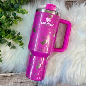 May include: A bright pink Stanley tumbler with a handle and a clear lid featuring a pink top. The exterior is decorated with a repeating pattern of silver-toned bows and small hearts. The Stanley logo is printed in white on the upper section.