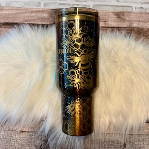 May include: A black and gold tumbler with a bee and honeycomb design. The tumbler has the name "Anna" in gold script. The design includes bees, flowers, and honeycomb patterns. The tumbler is approximately 10 inches tall.