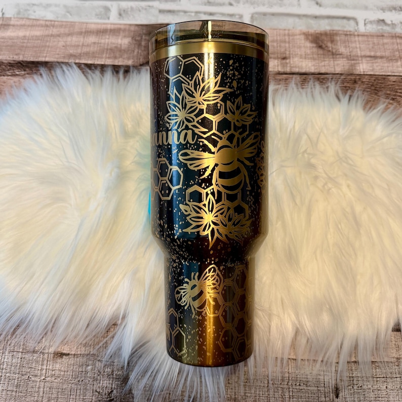 May include: A black and gold tumbler with a bee and honeycomb design. The tumbler has the name "Anna" in gold script. The design includes bees, flowers, and honeycomb patterns. The tumbler is approximately 10 inches tall.
