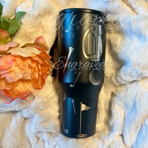 A black tumbler with golf-themed designs, including a golf club, ball, and flag, is placed on a white surface next to a pink flower.