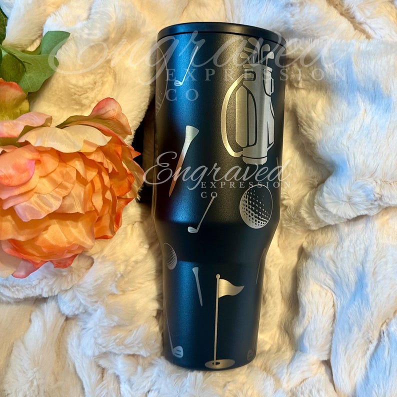 A black tumbler with golf-themed designs, including a golf club, ball, and flag, is placed on a white surface next to a pink flower.
