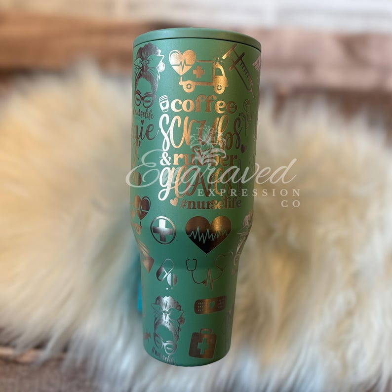 May include: A green stainless steel tumbler with gold-colored engraved designs. The tumbler features medical-themed graphics, including hearts, a cross, a stethoscope, and the words "coffee, scrubs, & rubber gloves." The text "Engraved Expression Co" is also visible.