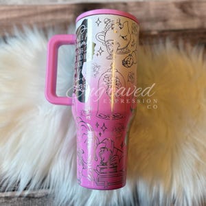 A pink and silver tumbler mug with a handle, featuring various Disney-themed illustrations and text.