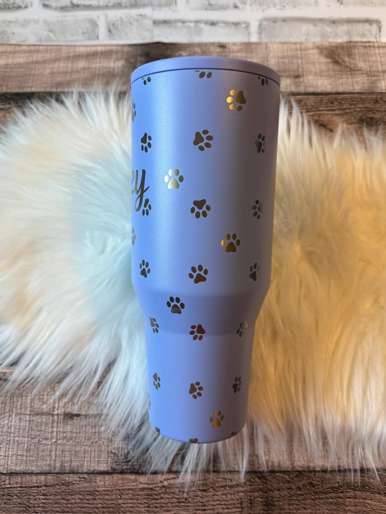 A blue tumbler with paw prints printed on it, placed on a white furry surface.