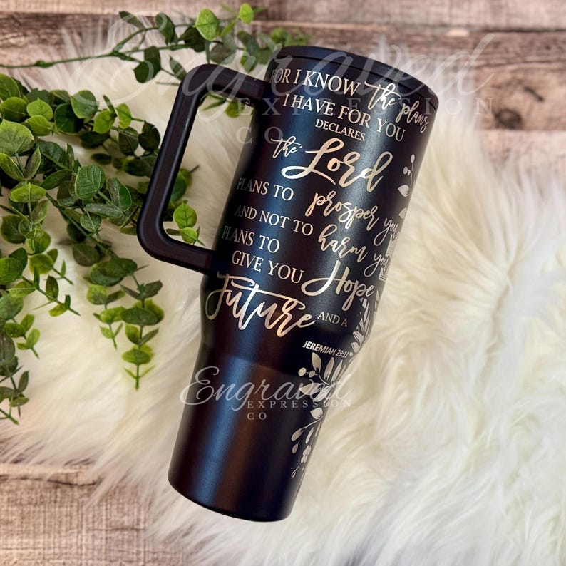 A black insulated tumbler mug with text printed on it, placed on a white fur rug.