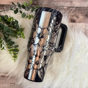 A black and silver floral patterned tumbler mug is placed on a white fur rug, with a plant branch visible to the left.