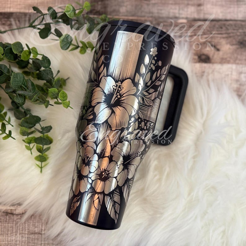A black and silver floral patterned tumbler mug is placed on a white fur rug, with a plant branch visible to the left.