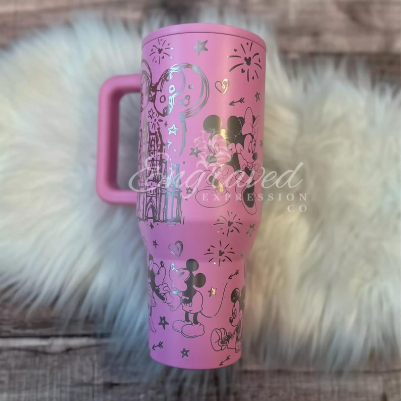 A pink tumbler mug with a handle and various Disney-themed designs, including Minnie Mouse and Mickey Mouse, printed on it.