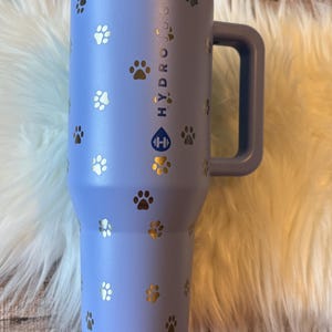 A blue insulated travel mug with paw prints printed on it, placed on a white fur rug.