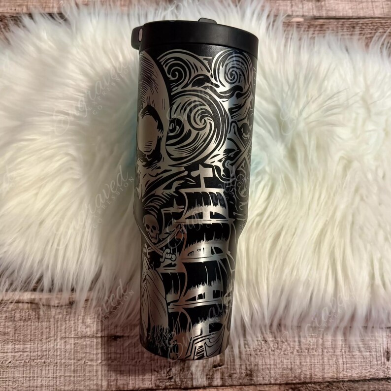 A black and silver tumbler mug with a skull and crossbones design on it, placed on a white furry surface.