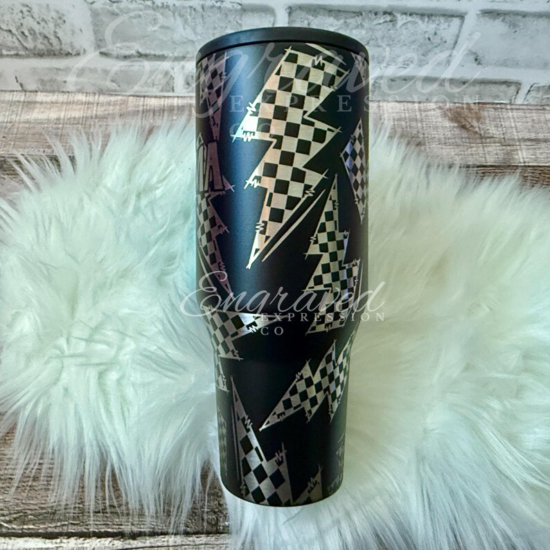 A black and white checkered patterned tumbler sits on a white furry surface, with a brick wall in the background.