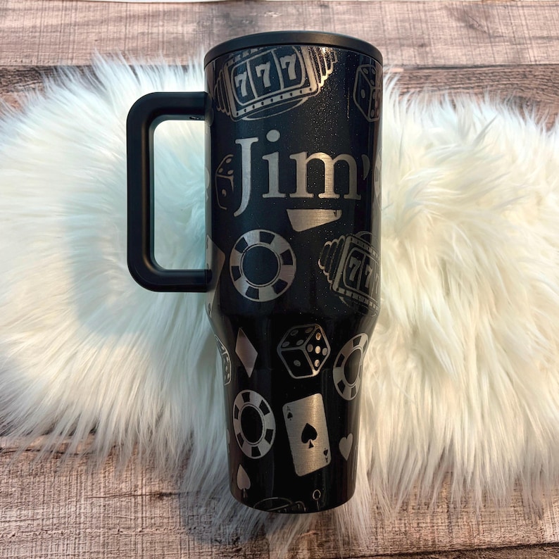 May include: Black tumbler with a handle, decorated with casino-themed graphics including dice, poker chips, and a slot machine. The name "Jim" is in silver lettering. The tumbler is designed for hot or cold drinks.