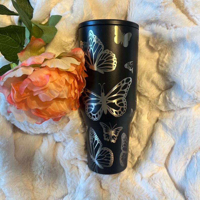 May include: A black tumbler with silver butterfly designs. The stainless steel tumbler has a matte finish and features various butterfly silhouettes. A large, orange and pink flower is next to the tumbler.
