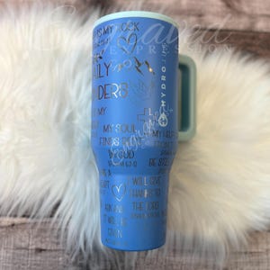 A blue tumbler mug with various religious and inspirational phrases printed on it, placed on a white fur surface.