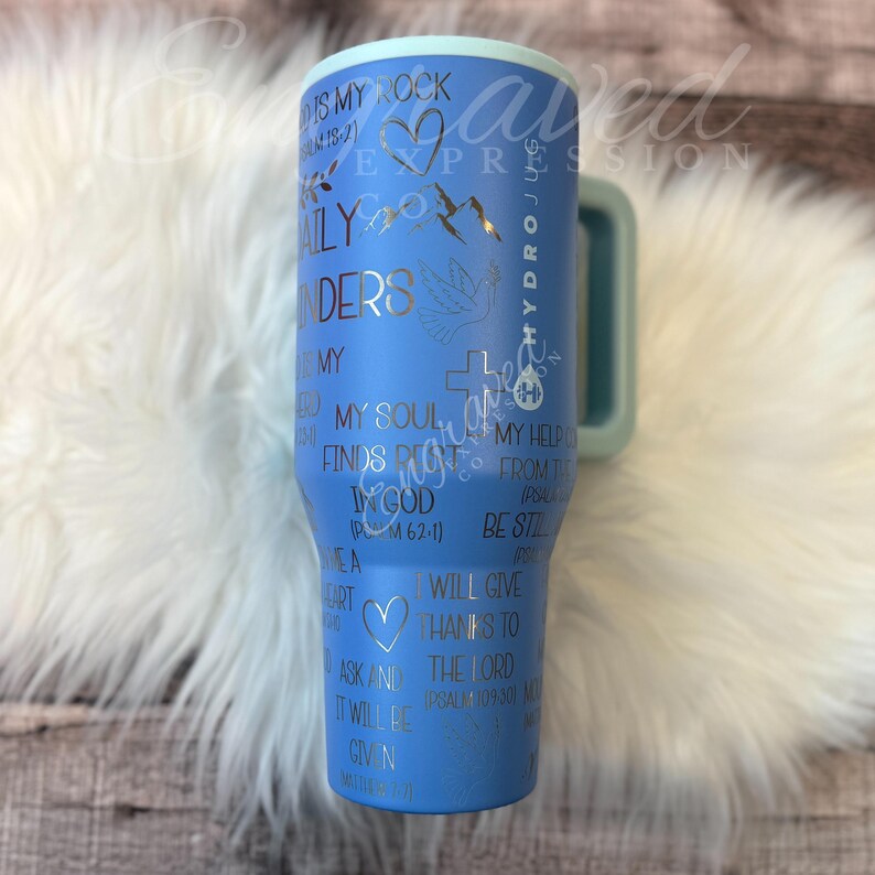 A blue tumbler mug with various religious and inspirational phrases printed on it, placed on a white fur surface.