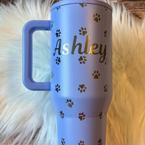 A personalized purple tumbler mug with the name "Ashley" printed on it, featuring paw prints.