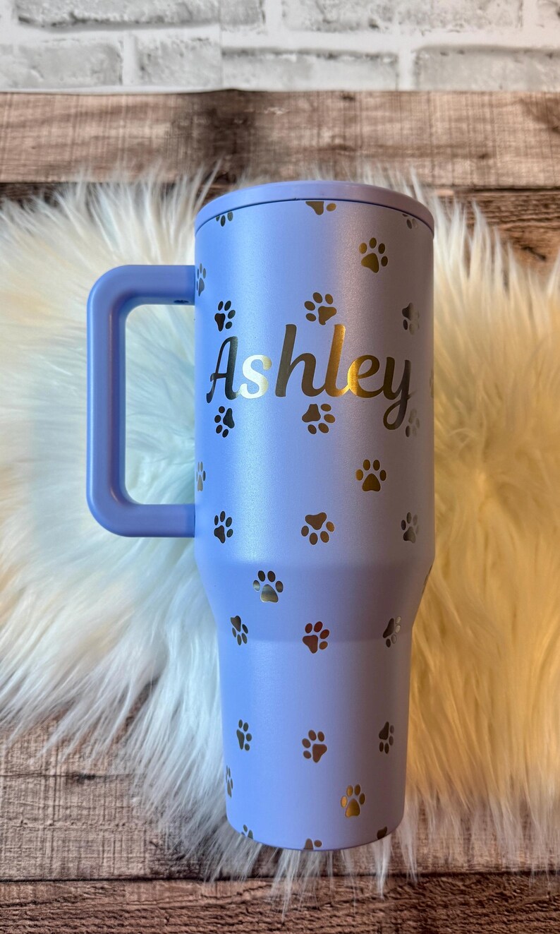 A personalized purple tumbler mug with the name "Ashley" printed on it, featuring paw prints.