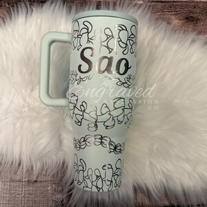 A turquoise insulated tumbler mug with a handle and a personalized name printed on it, placed on a white furry surface.