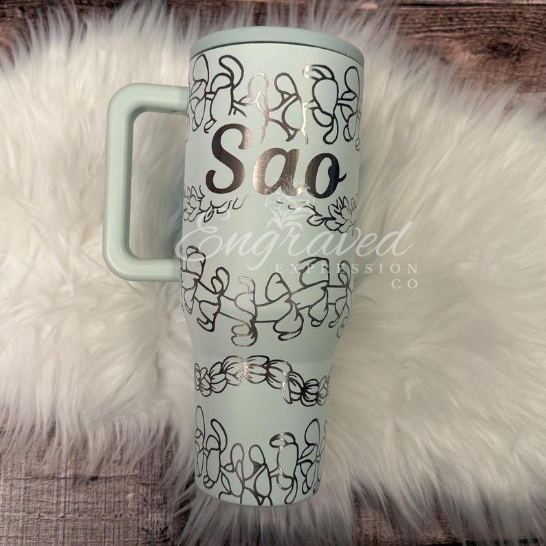 A turquoise insulated tumbler mug with a handle and a personalized name printed on it, placed on a white furry surface.
