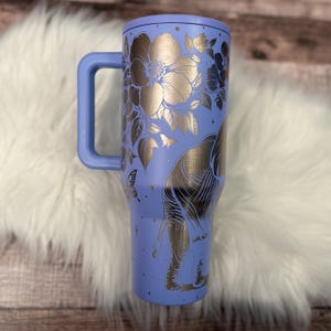 A blue insulated tumbler mug with a floral design and a handle, placed on a white fur rug.