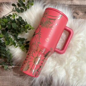 May include: A coral-colored Hydro Jug with a handle and a lid. The jug is adorned with a gold floral design, including daisies and other botanical elements. The brand name "HYDRO JUG" is visible on the side.