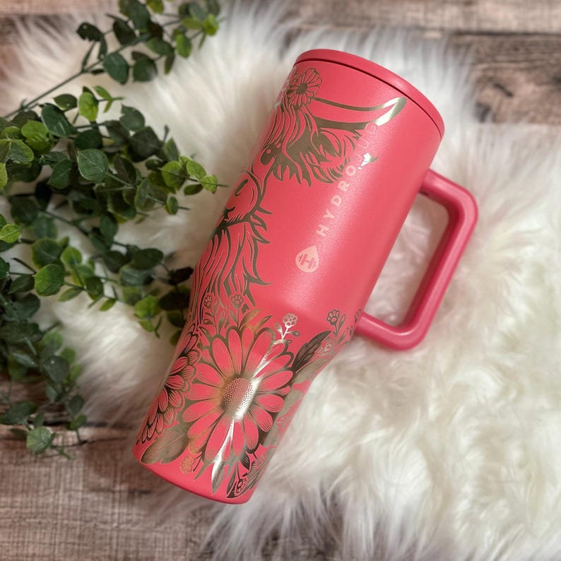 May include: A coral-colored Hydro Jug with a handle and a lid. The jug is adorned with a gold floral design, including daisies and other botanical elements. The brand name "HYDRO JUG" is visible on the side.