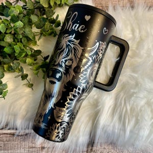 May include: Black stainless steel tumbler with a handle, featuring a silver engraved Highland cow design and sunflowers. The name "Shae" is also engraved. The tumbler has a black lid and handle, and is set against a white fur background.