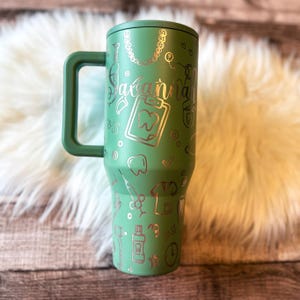 A green tumbler mug with various dental-related icons and text printed on it, placed on a white fur rug.