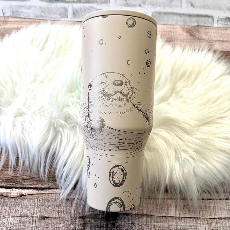 A white ceramic mug with a black and white illustration of an otter or seal on it, placed on a white fur rug.