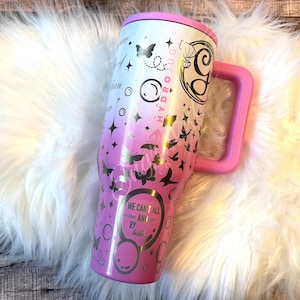 A pink and white tumbler mug with various designs and text printed on it, placed on a white furry surface.