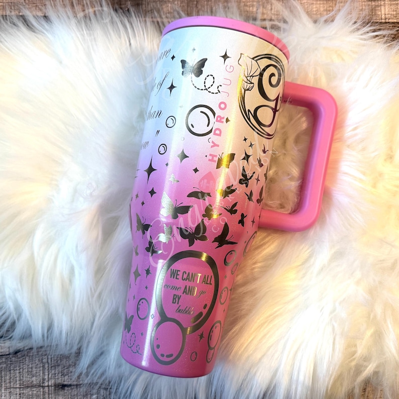A pink and white tumbler mug with various designs and text printed on it, placed on a white furry surface.