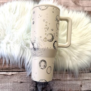 A white and beige insulated travel mug with a handle, placed on a white furry surface.