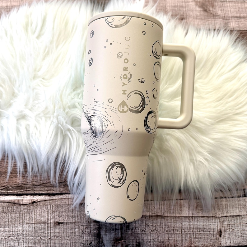 A white and beige insulated travel mug with a handle, placed on a white furry surface.
