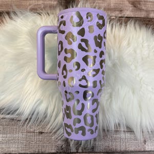 A purple insulated tumbler mug with a leopard print design is placed on a white furry surface.