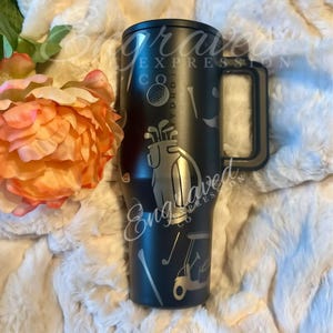 A black tumbler mug with golf-themed graphics and a handle, placed on a white fur-lined surface next to a pink flower.