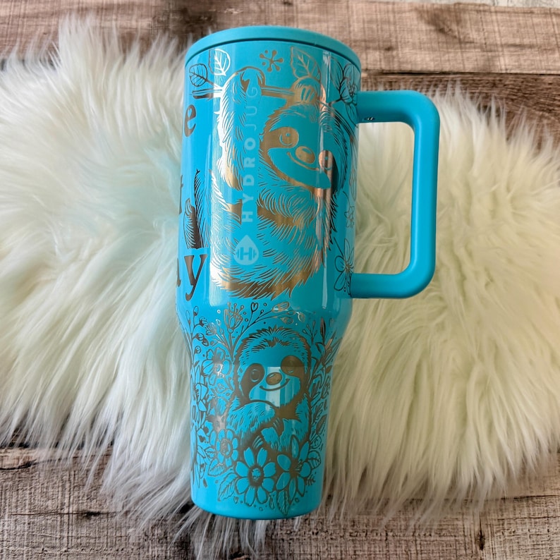 May include: A turquoise Hydrojug water bottle with a handle and a lid. The bottle features gold illustrations of sloths, leaves, and flowers. The text "Hydrojug" and "be the lazy" are also visible on the bottle.