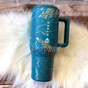 Puede incluir: Vaso Hydrojug turquesa con asa, con dise&ntilde;os dorados grabados de un delf&iacute;n, peces y coral. El vaso tiene el texto "HYDROJUG" y "Engraved Expression Co."