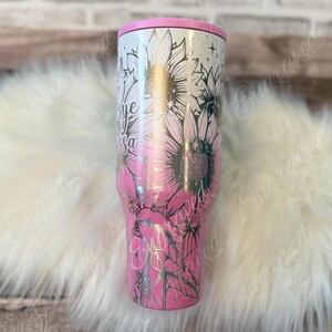 A pink and white tumbler with a floral design, including sunflowers and leaves, is displayed on a white fur surface.