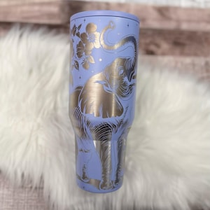 A purple insulated tumbler with a gold elephant design on it, placed on a white fur surface.
