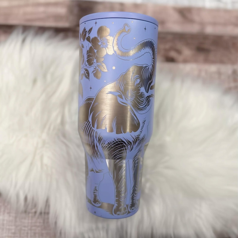 A purple insulated tumbler with a gold elephant design on it, placed on a white fur surface.
