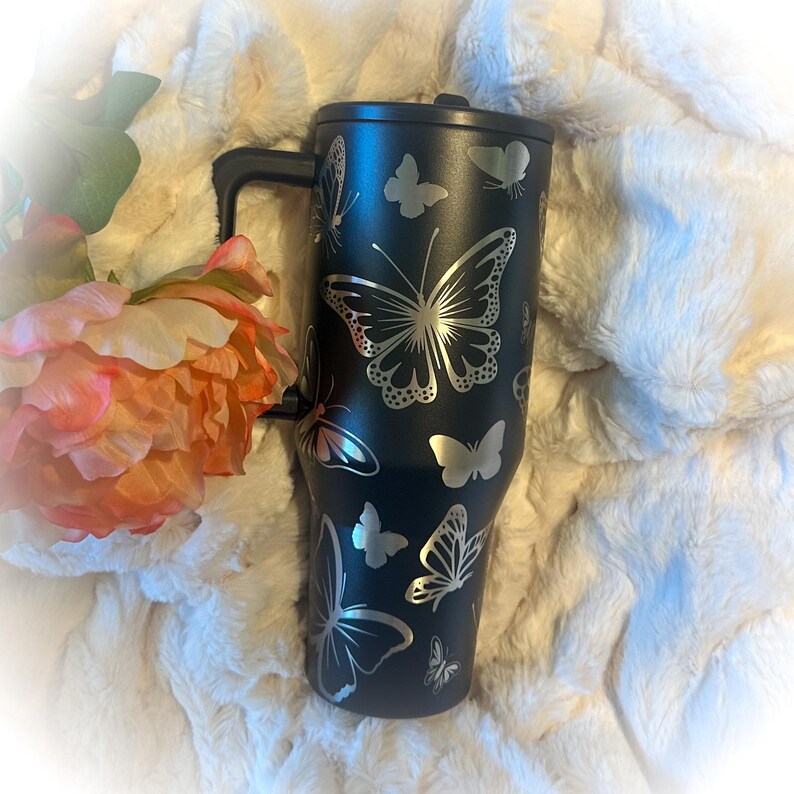 May include: A black tumbler with a handle, adorned with silver butterfly designs. The tumbler is set against a soft, white, fluffy background, with a pink and orange flower in the corner.