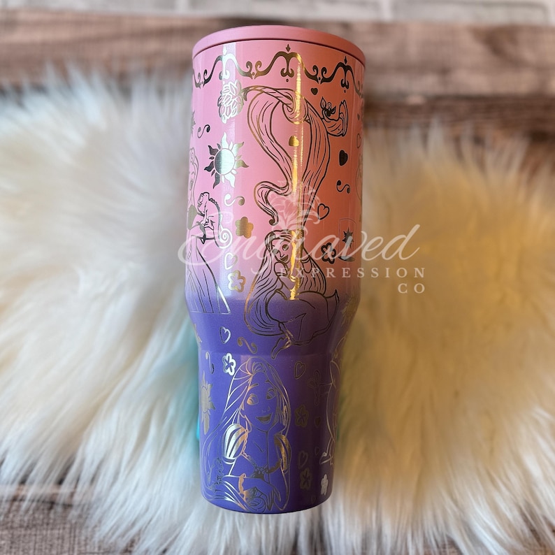 A pink and purple tumbler with a Disney princess design, featuring a princess and a unicorn, is shown against a white fur background.