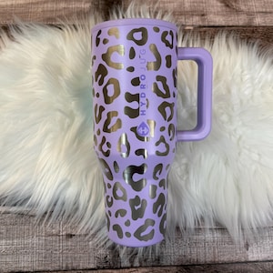 A purple and black leopard print thermos mug with the text "HYDRO JUG" printed on it, placed on a white fur rug.