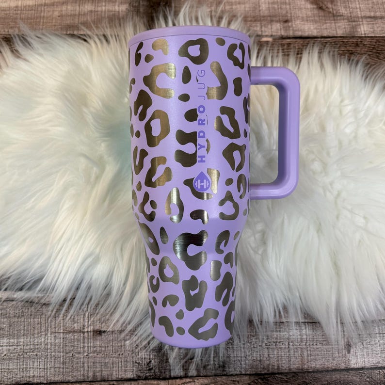 A purple and black leopard print thermos mug with the text "HYDRO JUG" printed on it, placed on a white fur rug.