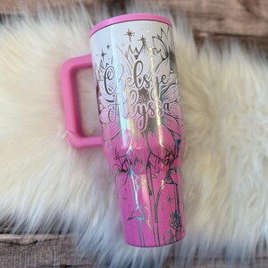 A pink and white tumbler mug with a floral design and text printed on it, placed on a white furry surface.