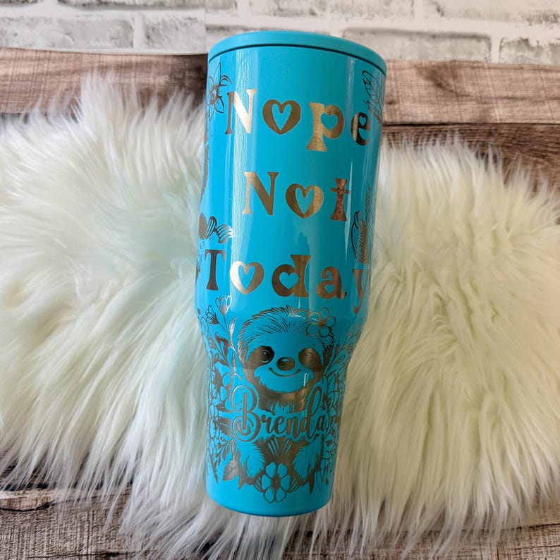 May include: A turquoise tumbler with a gold design and the words "Nope Not Today." The design includes a sloth and floral accents. The name "Brenda" is also featured. The tumbler is on a white fluffy surface.